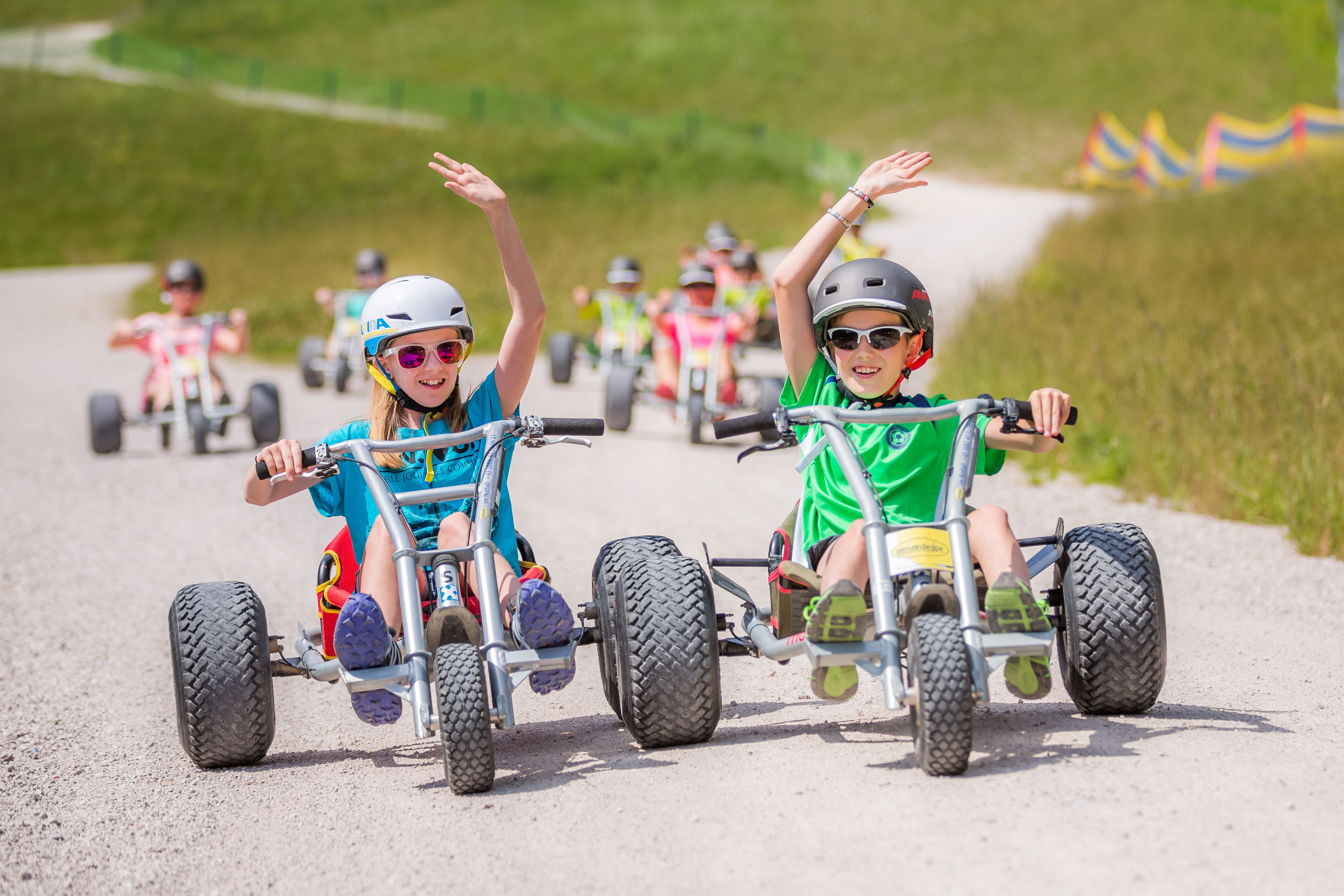 Zwei Kinder fahren nebeneinander in Mountaincarts und heben jeweils eine Hand; weitere Personen folgen ihnen.