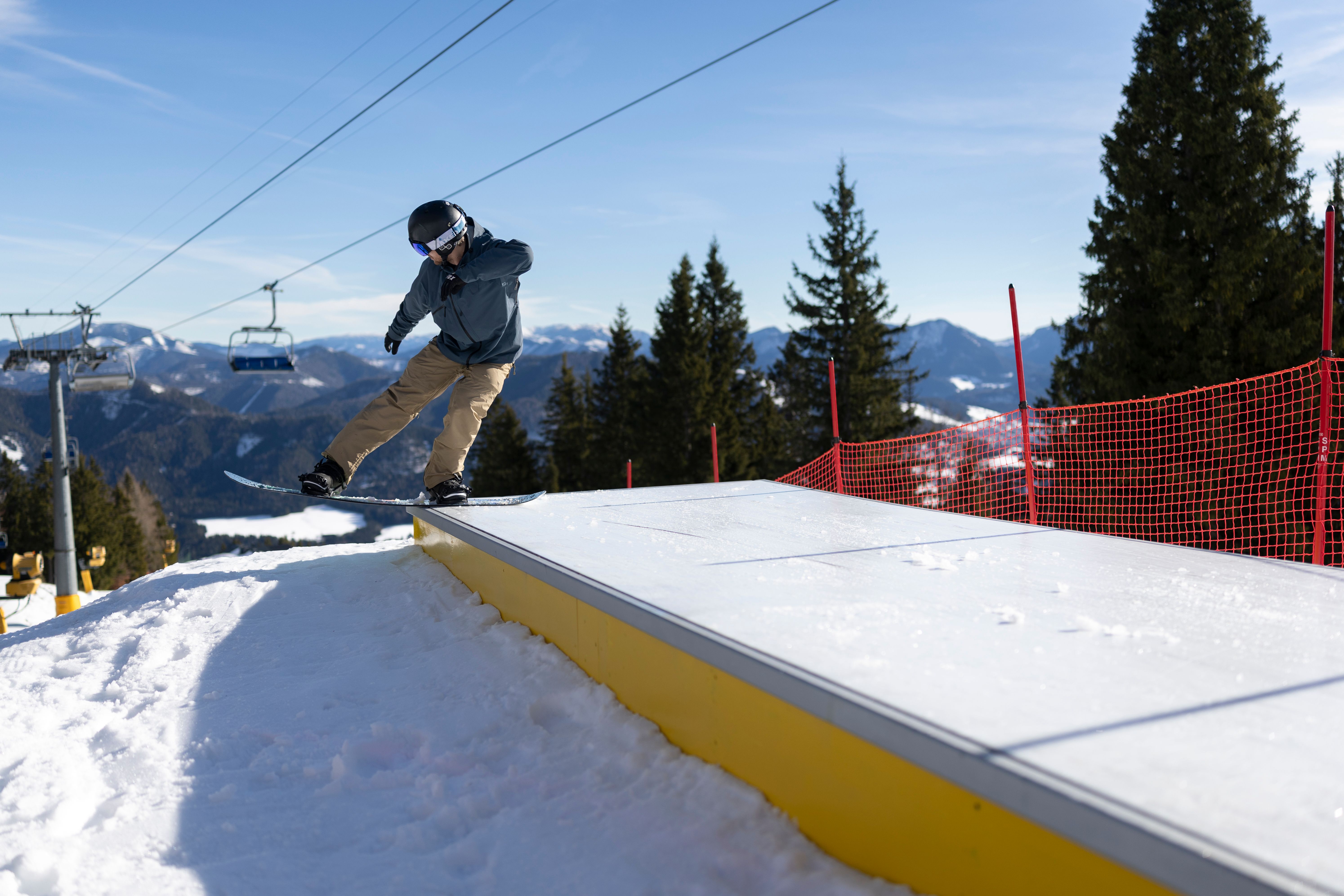 Snowboarder fährt über ein breites Hindernis im Snowpark; im Hintergrund verläuft ein Sessellift.