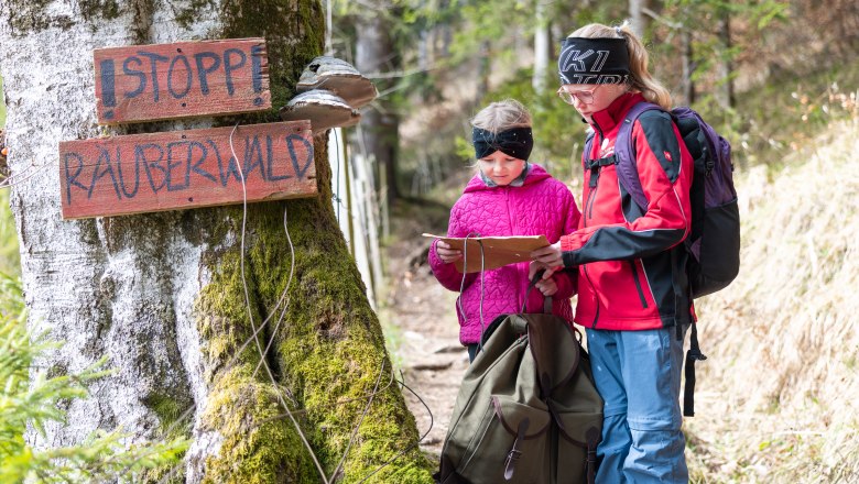 Zwei Kinder stehen auf einem Waldweg neben einem moosbewachsenen Baum mit einem Holzschild &bdquo;STOPP R&Auml;UBERWALD&ldquo;. Die beiden tragen Outdoor-Kleidung und Rucks&auml;cke und schauen gemeinsam auf eine Karte. Im Hintergrund ist ein heller Mischwald zu sehen.