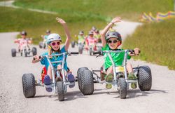 Zwei Kinder fahren nebeneinander in Mountaincarts und heben jeweils eine Hand; weitere Personen folgen ihnen.