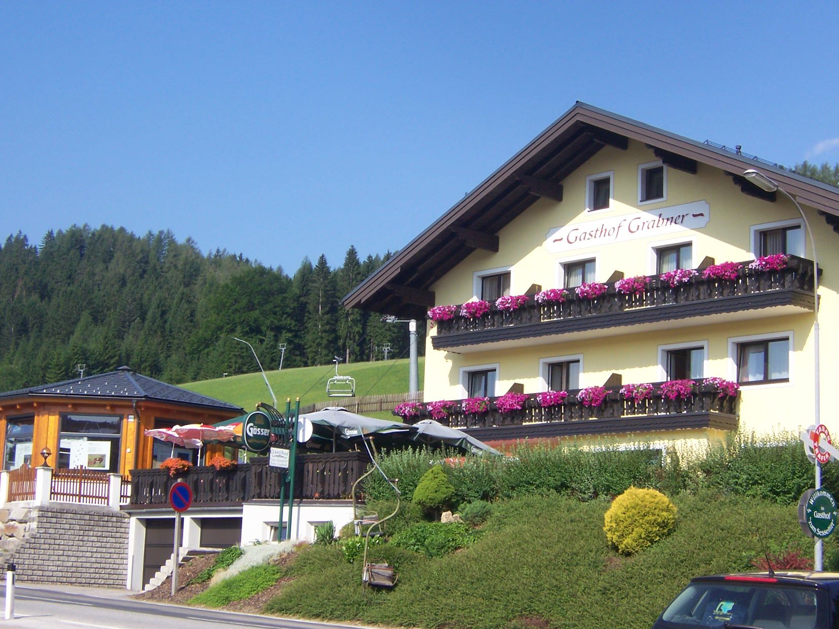 Gasthof Grabner mit blumengeschmückten Balkonen, Gastgarten und Sessellift am grünen Hang.