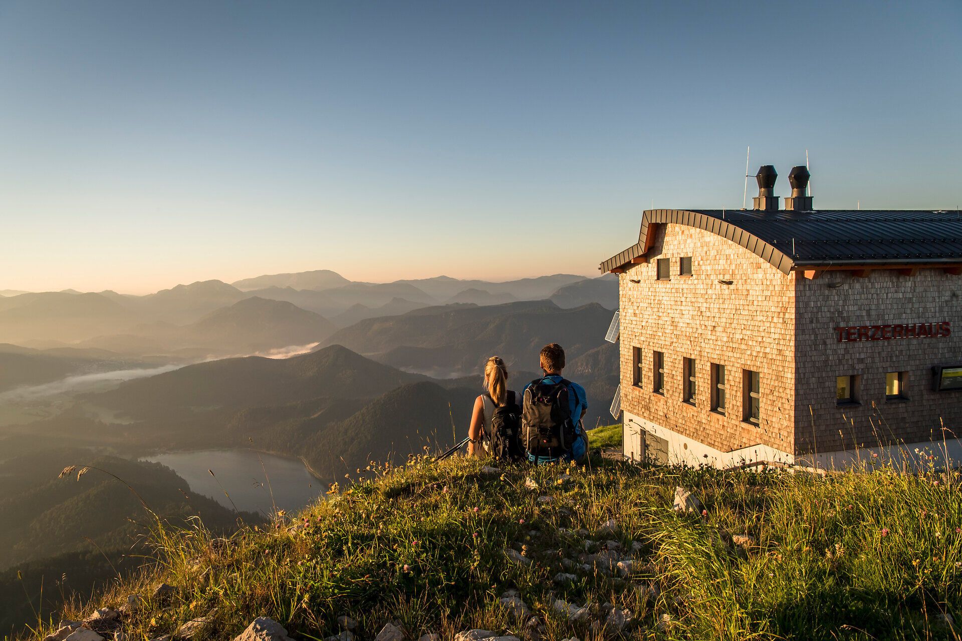 Die sanften Hügel und majestätischen Berge umarmen die Szenerie, während die warmen Sonnenstrahlen den Tag erhellen. Ein Paar genießt den atemberaubenden Ausblick und die friedliche Atmosphäre, die nur die Natur bieten kann. Hier, wo die Zeit stillzustehen scheint, wird jeder Moment zu einem unvergesslichen Erlebnis.