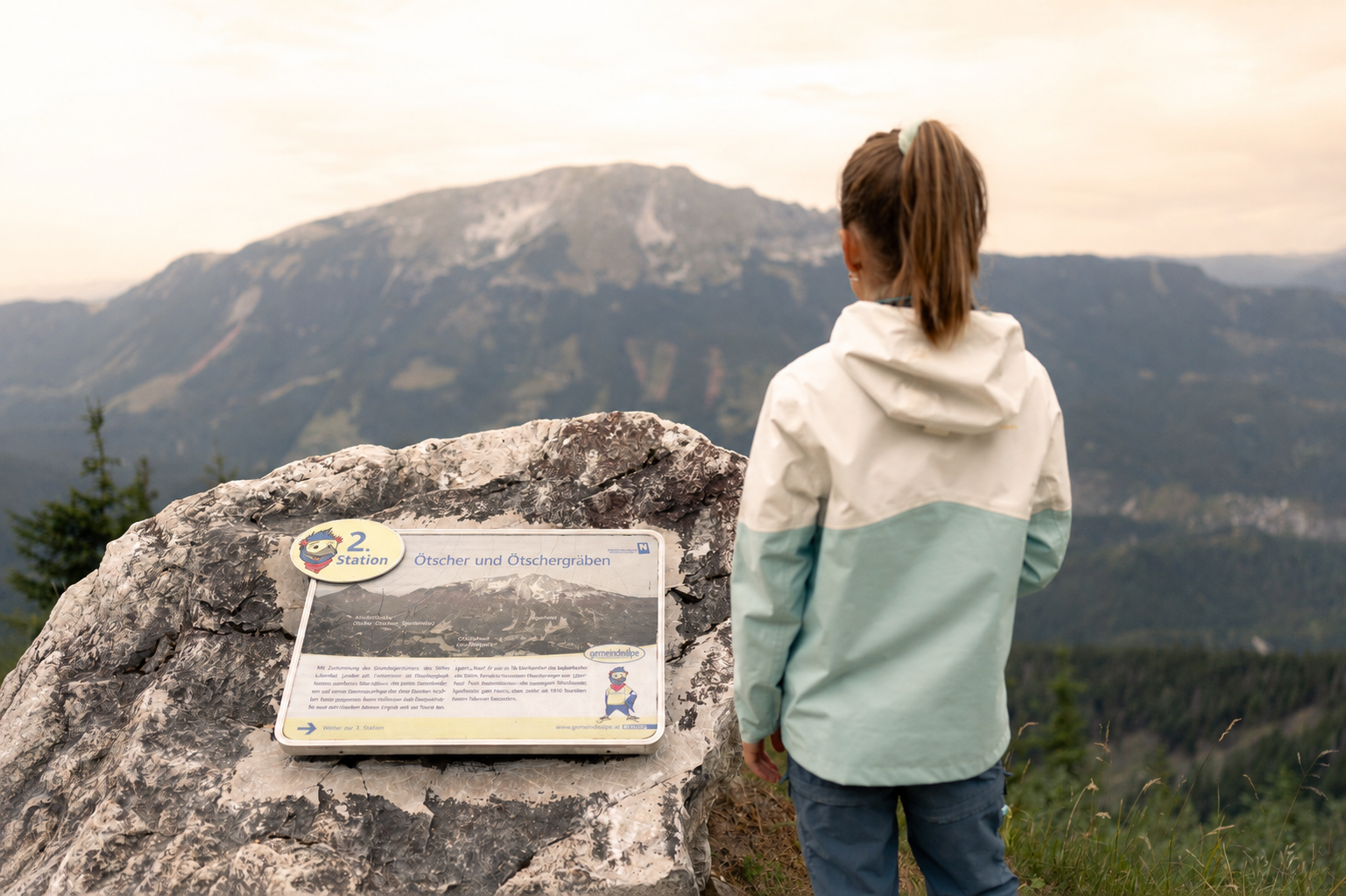 Mädchen steht neben einer Infotafel auf einem Felsen und blickt auf den Ötscher unter warmem, hellem Himmel.