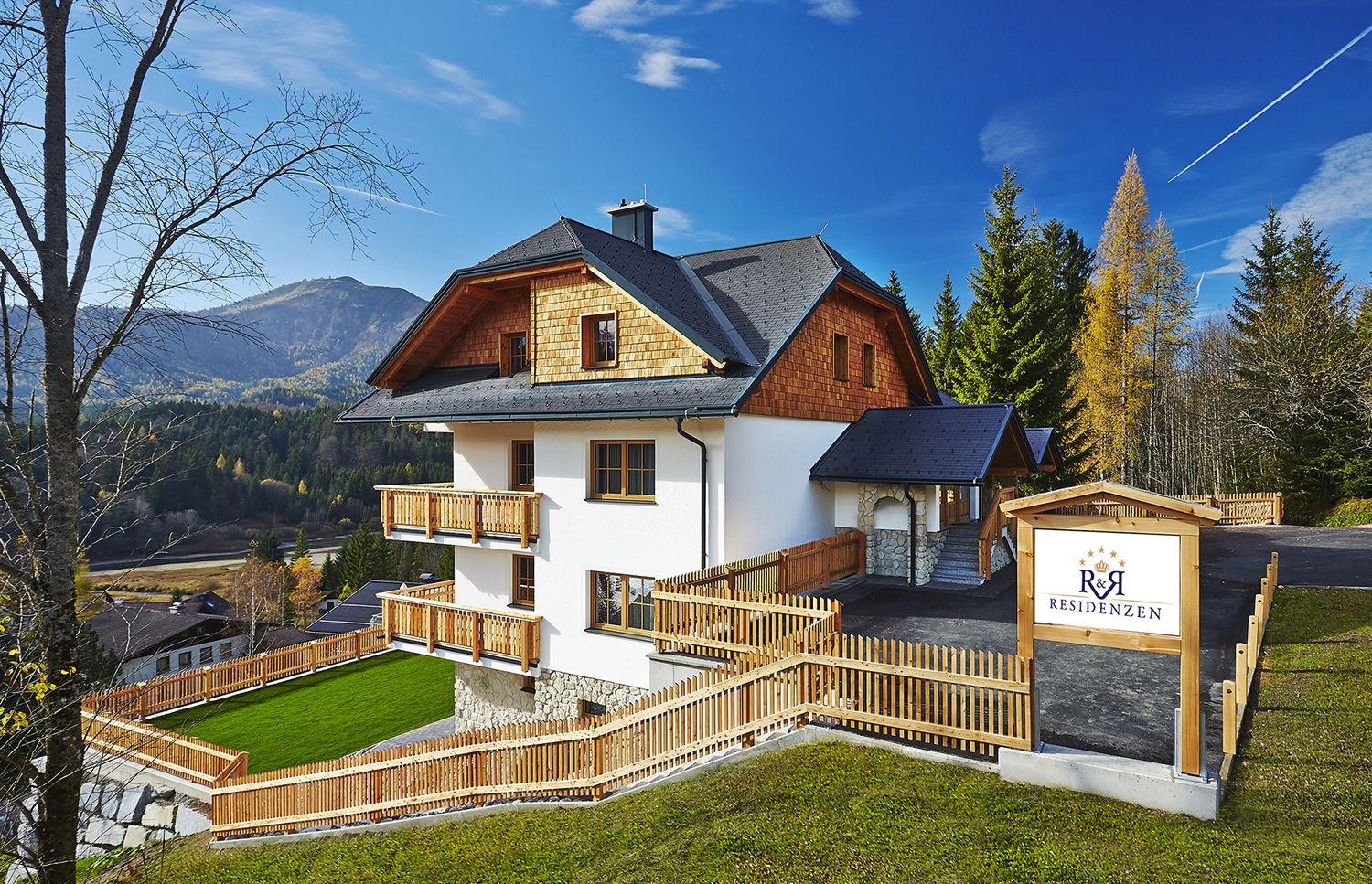 Ein modernes Haus in alpiner Umgebung mit Holzverkleidung und Balkonen, umgeben von Bäumen und Bergen. Ein Schild zeigt 'R&R Residenzen'.