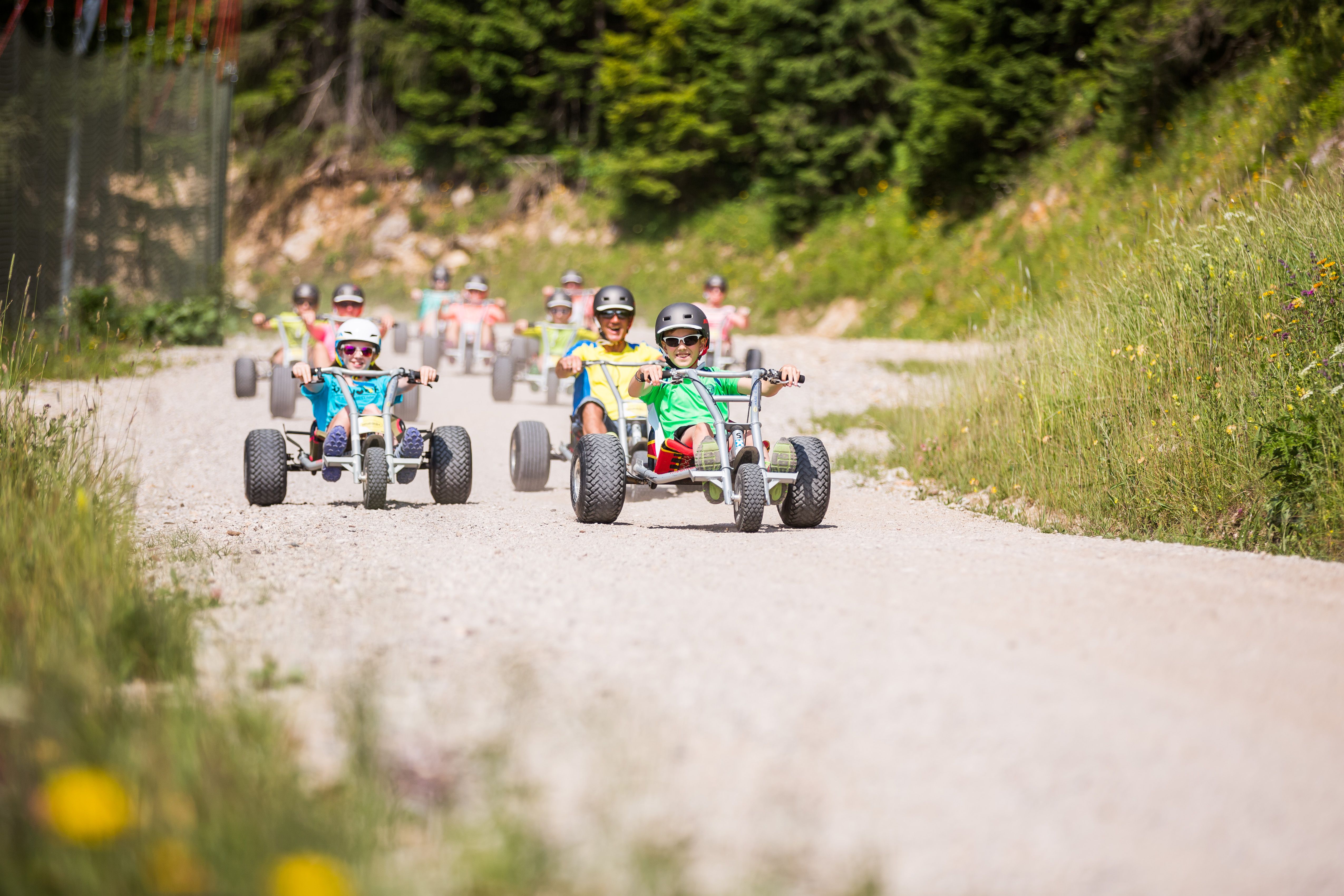 Kinder und Erwachsene fahren mit Helmen in Mountaincarts eine Schotterstraße hinunter.
