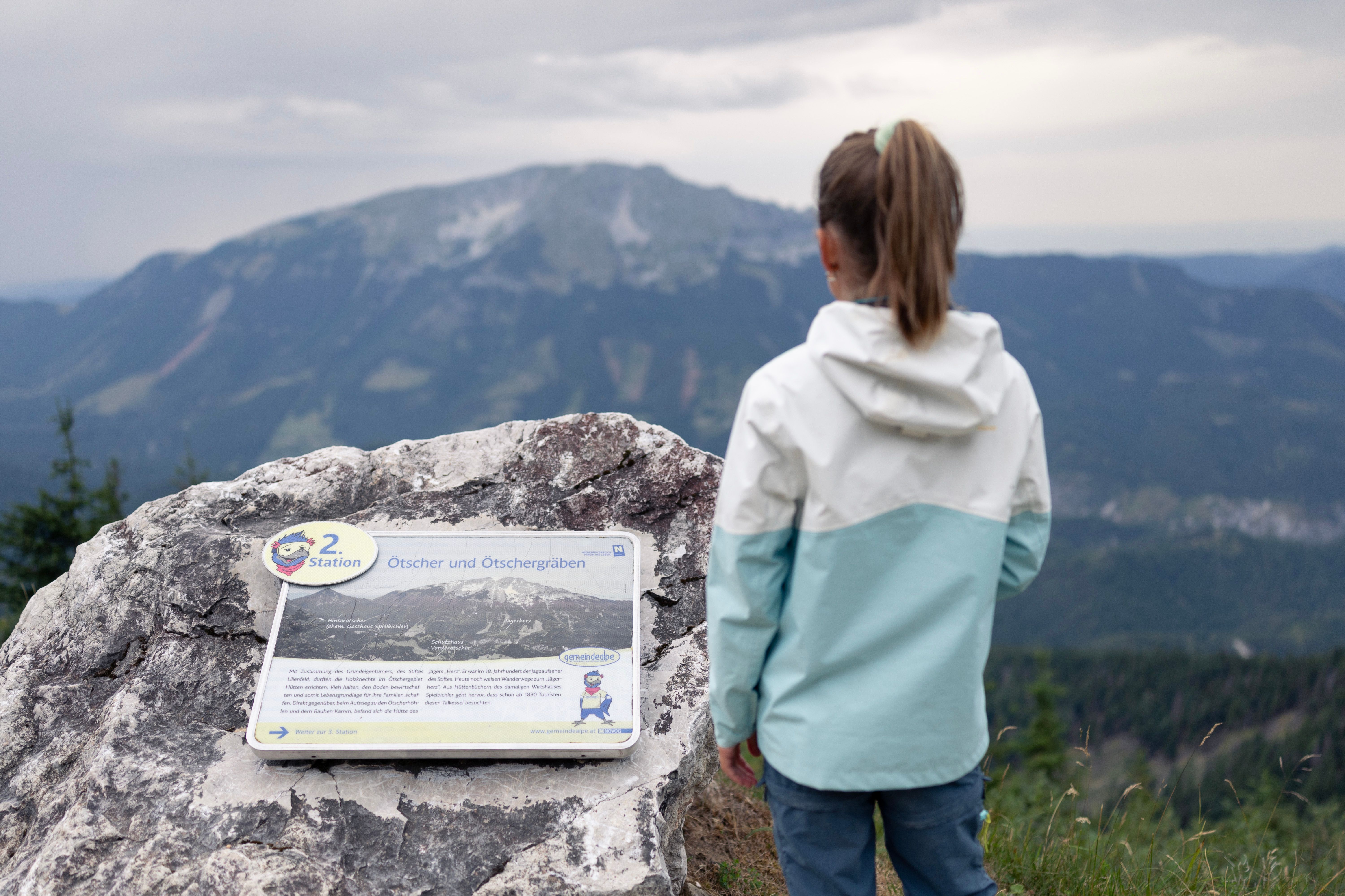 Mädchen betrachtet neben einer Infotafel den Ötscher und die umliegende Berglandschaft.