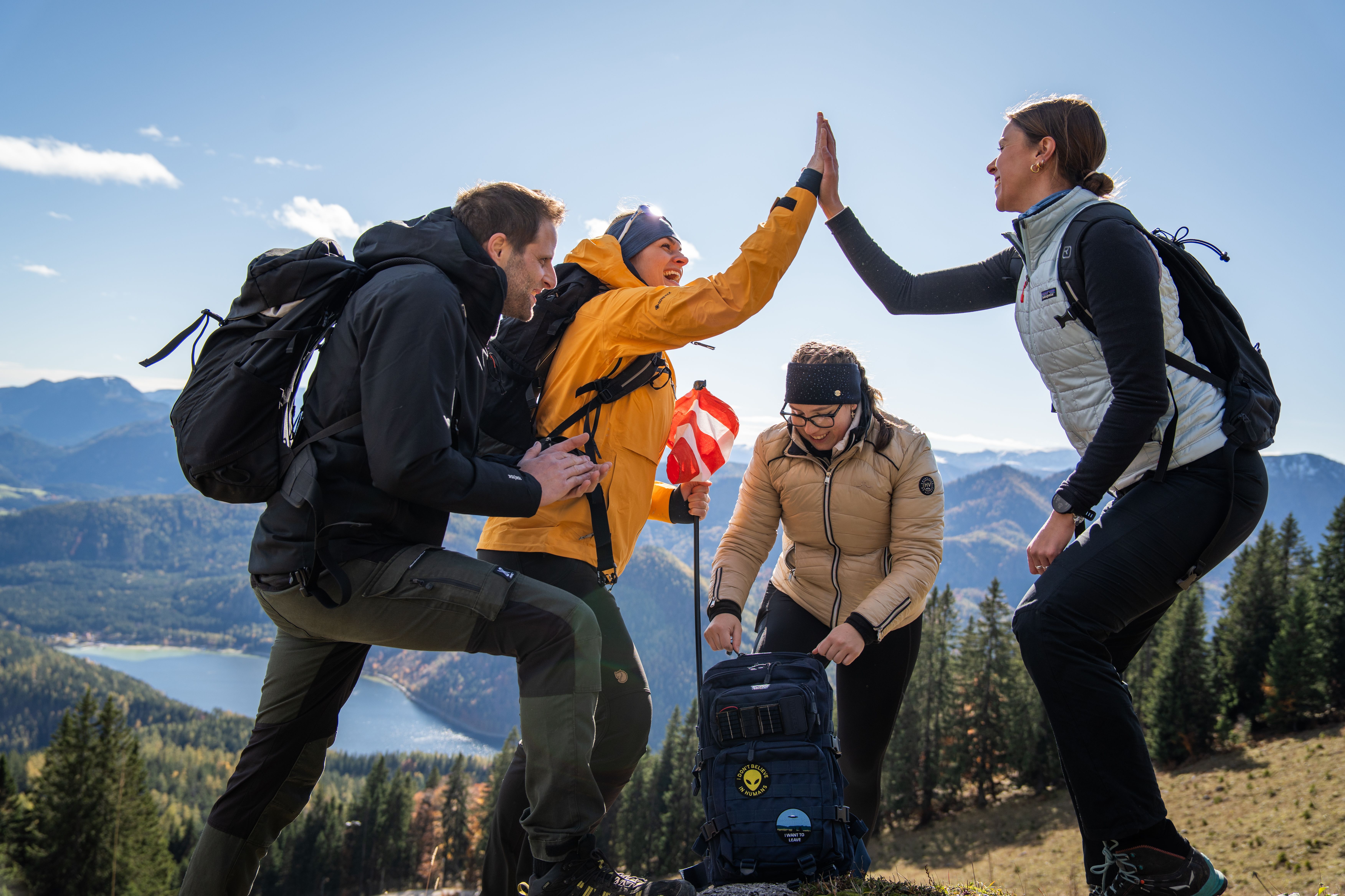 Vier Wandernde feiern gemeinsam mit einem High-Five vor Berg- und Seekulisse.