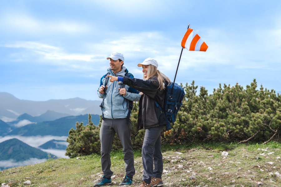 Zwei Wanderer mit Rucksäcken und Mütze auf einem Berg, eine zeigt in die Ferne, im Hintergrund Berglandschaft und Wolken