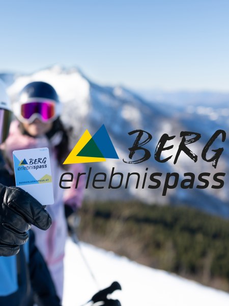 Zwei Personen in Skiausr&uuml;stung vor verschneiten Bergen; die vordere Person h&auml;lt den Bergerlebnispass in die Kamera.
