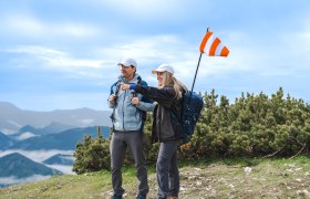 Zwei Wanderer mit Rucksäcken und Mütze auf einem Berg, eine zeigt in die Ferne, im Hintergrund Berglandschaft und Wolken
