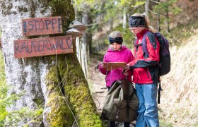 Zwei Kinder stehen auf einem Waldweg neben einem moosbewachsenen Baum mit einem Holzschild &bdquo;STOPP R&Auml;UBERWALD&ldquo;. Die beiden tragen Outdoor-Kleidung und Rucks&auml;cke und schauen gemeinsam auf eine Karte. Im Hintergrund ist ein heller Mischwald zu sehen.