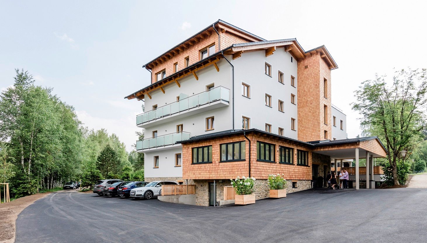 Ein modernes Hotelgebäude mit Holzverkleidung und Balkonen, umgeben von Bäumen und einem Parkplatz mit Autos.