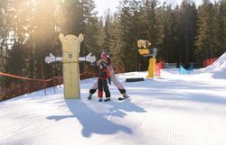 Erwachsene unterstützt ein Kind auf Ski neben einer bunten Figur in der verschneiten Funslope