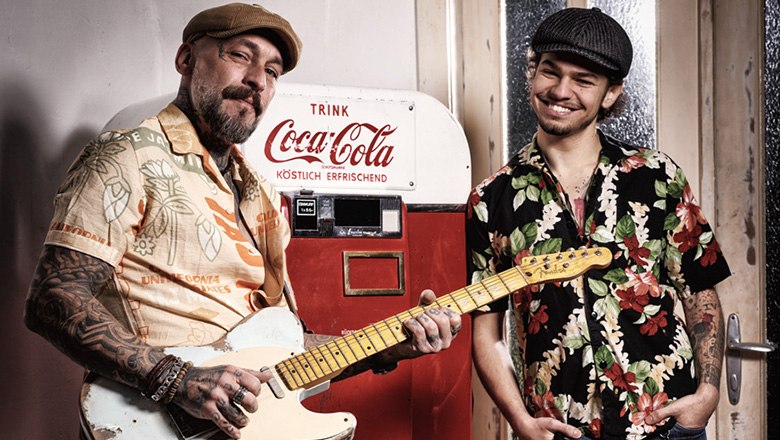 Zwei Musiker mit Retro-Look vor einem Coca-Cola-Automaten – einer spielt E-Gitarre, beide tragen bunte Hawaiihemden und Schiebermützen.