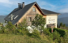 Holzhaus mit schwarzem Dach und Schriftzug 'Zum Bäreneck' in ländlicher Berglandschaft unter blauem Himmel