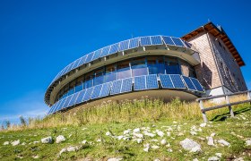 Panoramarestaurant Terzerhaus auf der Gemeindealpe Mitterbach mit moderner Glasfassade und Solarpaneelen, umgeben von Almwiese unter blauem Himmel.