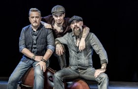 Drei M&auml;nner mit Tattoos und Armbanduhren sitzen eng zusammen vor schwarzem Hintergrund, zwei tragen gestreifte Hemden, einer eine Schieberm&uuml;tze