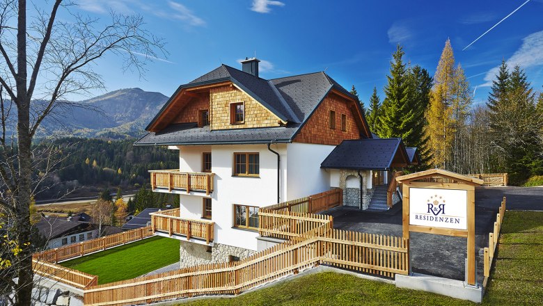 R&R Residenzen, © R&R Residenzen Ein modernes Haus in alpiner Umgebung mit Holzverkleidung und Balkonen, umgeben von Bäumen und Bergen. Ein Schild zeigt 'R&R Residenzen'.