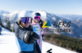 Zwei Personen in Skiausr&uuml;stung vor verschneiten Bergen; die vordere Person h&auml;lt den Bergerlebnispass in die Kamera.
