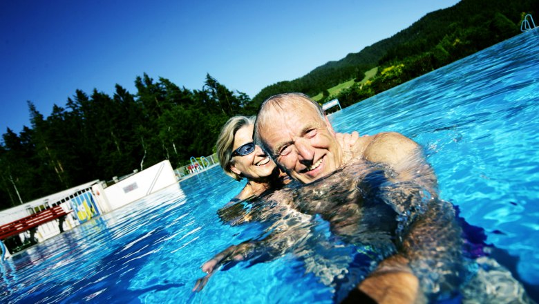 Ein &auml;lteres Paar schwimmt l&auml;chelnd in einem Freibad mit Wald im Hintergrund.