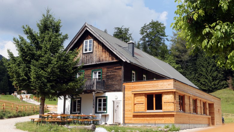 Ein traditionelles Holzhaus mit Anbau in einer l&auml;ndlichen Umgebung, umgeben von B&auml;umen und Wiesen.