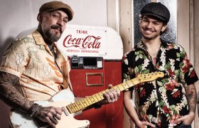 Zwei Musiker mit Retro-Look vor einem Coca-Cola-Automaten – einer spielt E-Gitarre, beide tragen bunte Hawaiihemden und Schiebermützen.