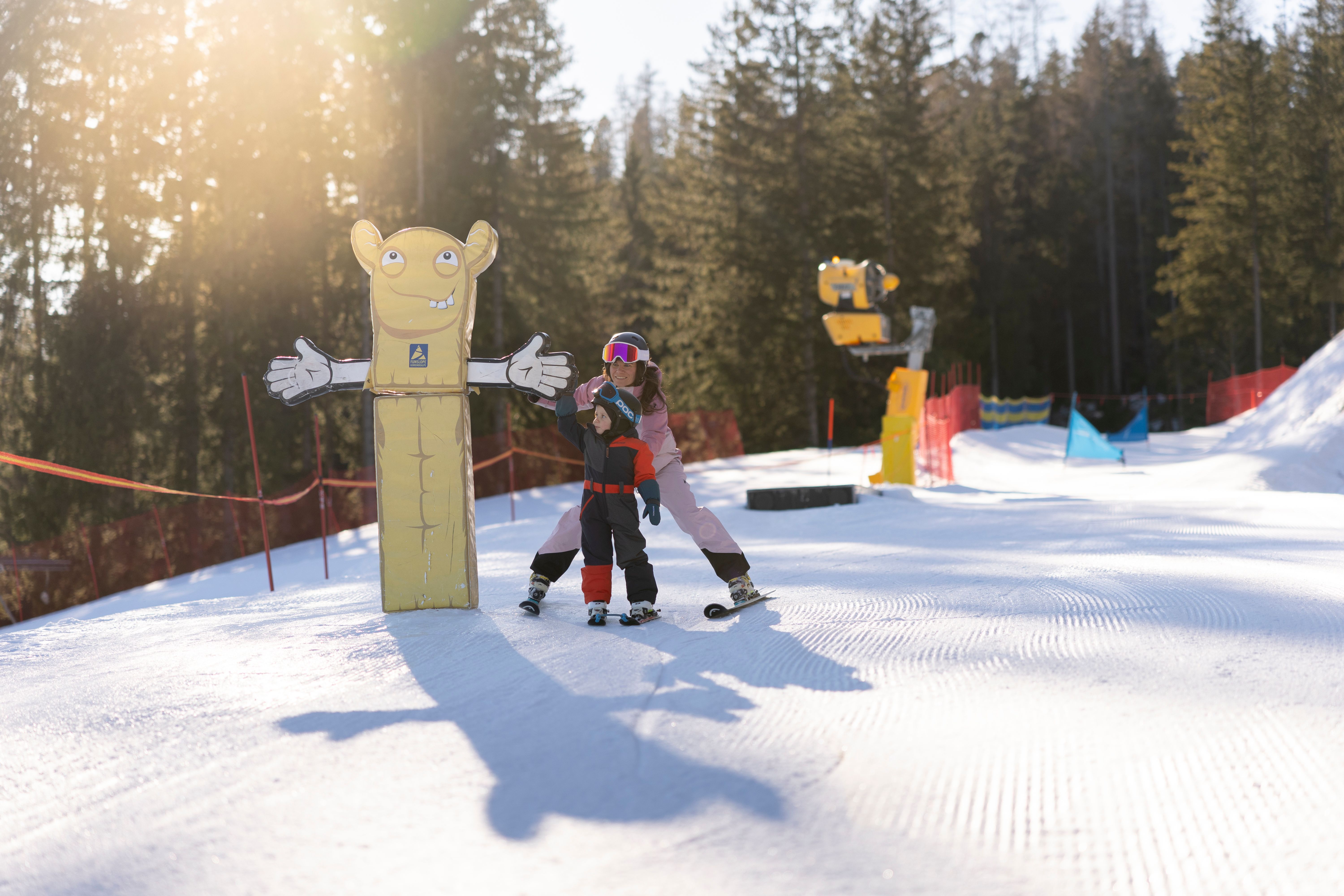 Erwachsene unterstützt ein Kind auf Ski neben einer bunten Figur in der verschneiten Funslope