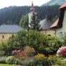 A picturesque garden with colorful flowers in front of traditional houses and a church in a mountainous landscape.