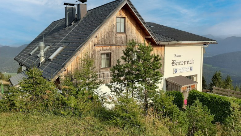 Holzhaus mit schwarzem Dach und Schriftzug 'Zum Bäreneck' in ländlicher Berglandschaft unter blauem Himmel
