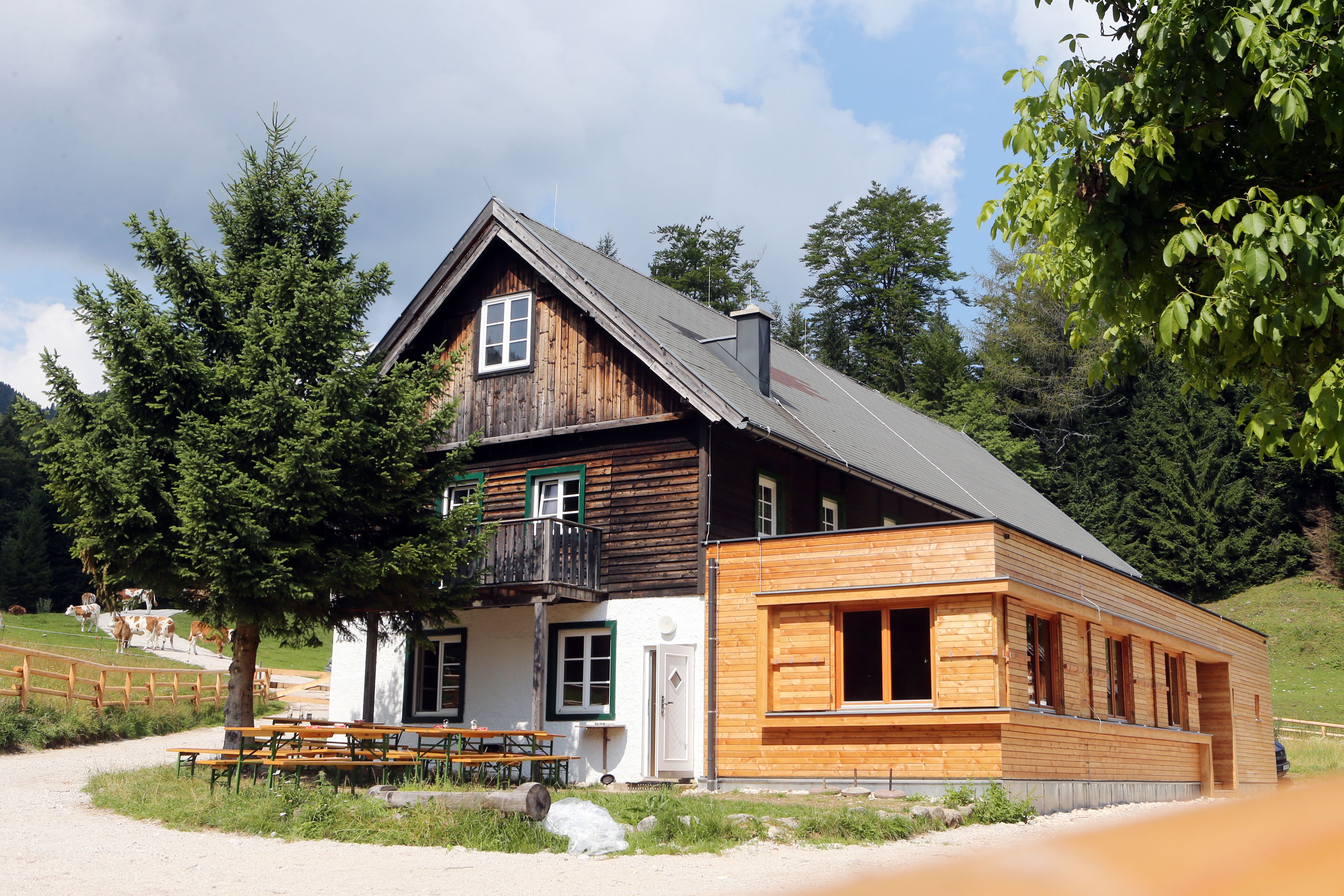 Ein traditionelles Holzhaus mit Anbau in einer ländlichen Umgebung, umgeben von Bäumen und Wiesen.